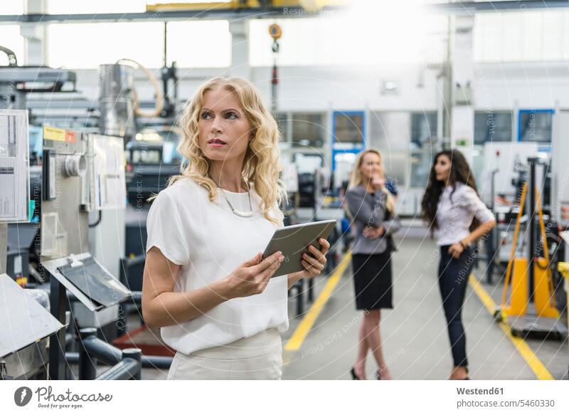 Frau hält Tablette in Fabrikhalle mit zwei Frauen im Hintergrund Tablet Computer Tablet-PC Tablet PC iPad Tablet-Computer Industriehallen Fabrikhallen Fabriken