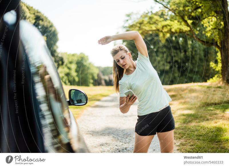 Sportliche junge Frau, die sich dehnt und ein Handy an einem Auto in einem Park benutzt Wagen PKWs Automobil Autos Parkanlagen Parks Mobiltelefon Handies Handys