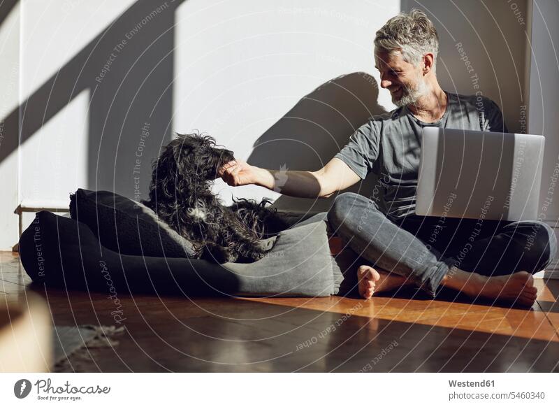 Reifer Mann sitzt zu Hause mit Laptop und Hund auf dem Boden Leute Menschen People Person Personen Alleinstehende Alleinstehender Singles Unverheiratete