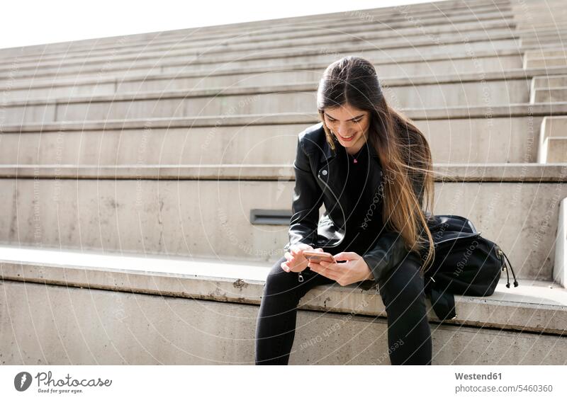 Lächelnde junge Frau sitzt auf einer Treppe und benutzt ein Mobiltelefon lächeln Treppenaufgang Handy Handies Handys Mobiltelefone weiblich Frauen sitzen