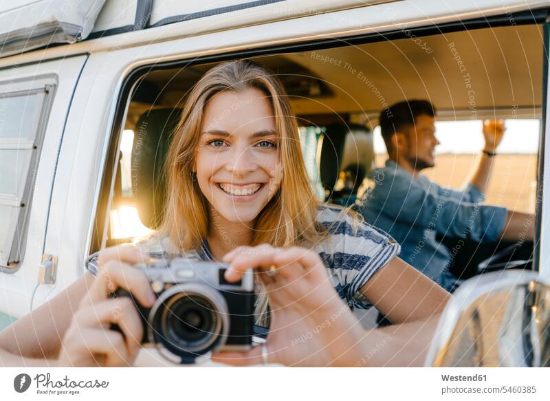 Porträt einer glücklichen Frau mit Kamera, die sich aus dem Fenster eines Wohnmobils lehnt, in dem ein Mann fährt Portrait Porträts Portraits Paar Pärchen Paare