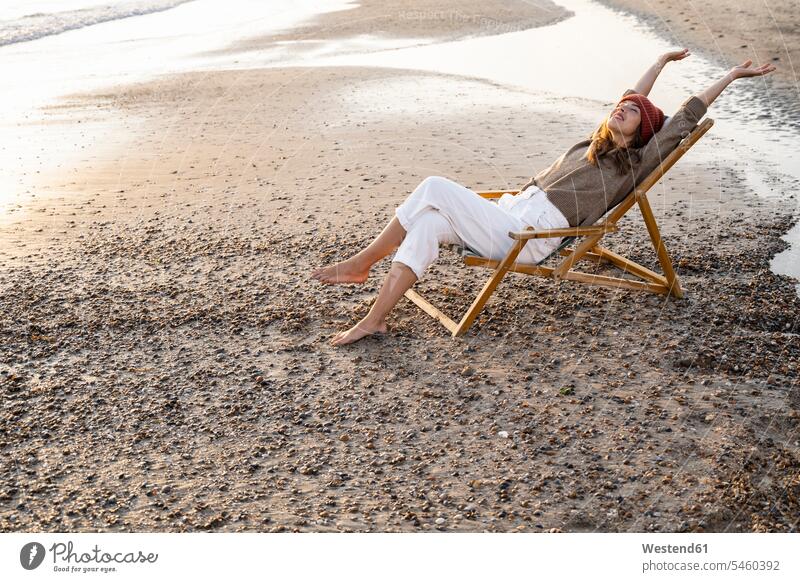 Lächelnde junge Frau sitzt bei Sonnenuntergang mit verschränkten Beinen und angehobenen Armen auf einem Klappstuhl am Strand Farbaufnahme Farbe Farbfoto