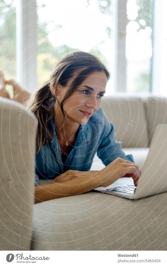 Lächelnde reife Frau liegt auf der Couch zu Hause mit Laptop weiblich Frauen Zuhause daheim liegen liegend lächeln Notebook Laptops Notebooks Sofa Couches Liege