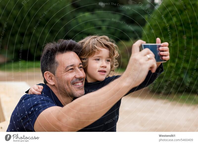 Glücklicher Vater und Sohn bei der Aufnahme eines Selfies im Park glücklich glücklich sein glücklichsein Parkanlagen Parks Papas Väter Vati Vatis Papis Söhne