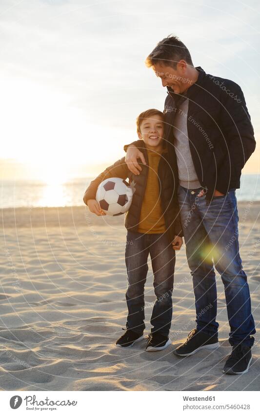 Vater umarmt Sohn mit Fussball am Strand bei Sonnenuntergang Beach Straende Strände Beaches Sonnenuntergänge umarmen Umarmung Umarmungen Arm umlegen Papas Väter