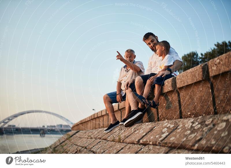 Großvater, Vater und Sohn sitzen auf einer Mauer am Flussufer Leute Menschen People Person Personen Europäisch Kaukasier kaukasisch Gruppe von Menschen