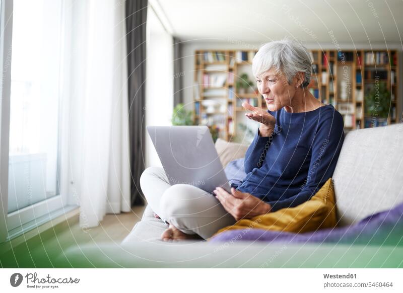 Ältere Frau gibt fliegenden Kuss bei Videoanruf, während sie zu Hause sitzt Farbaufnahme Farbe Farbfoto Farbphoto Innenaufnahme Innenaufnahmen innen drinnen Tag