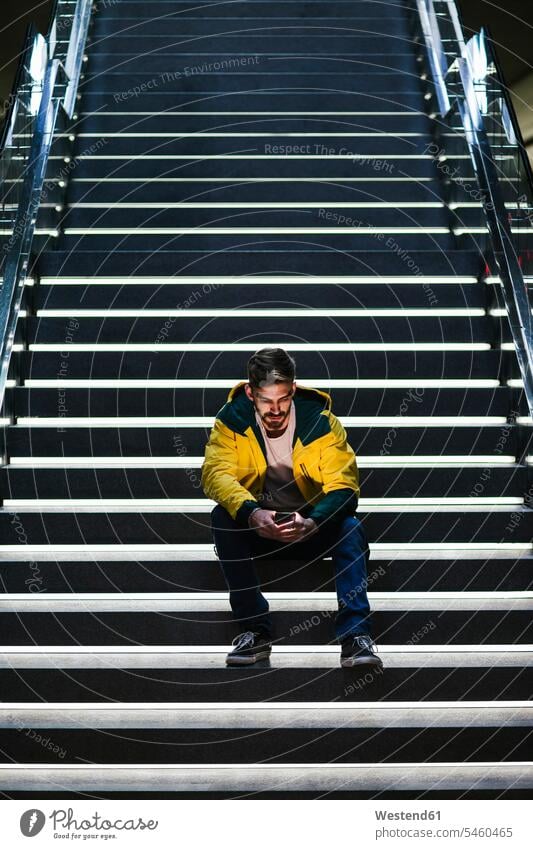 Mann sitzt auf einer Treppe in der U-Bahn-Station und benutzt ein Mobiltelefon Leute Menschen People Person Personen Europäisch Kaukasier kaukasisch 1 Ein
