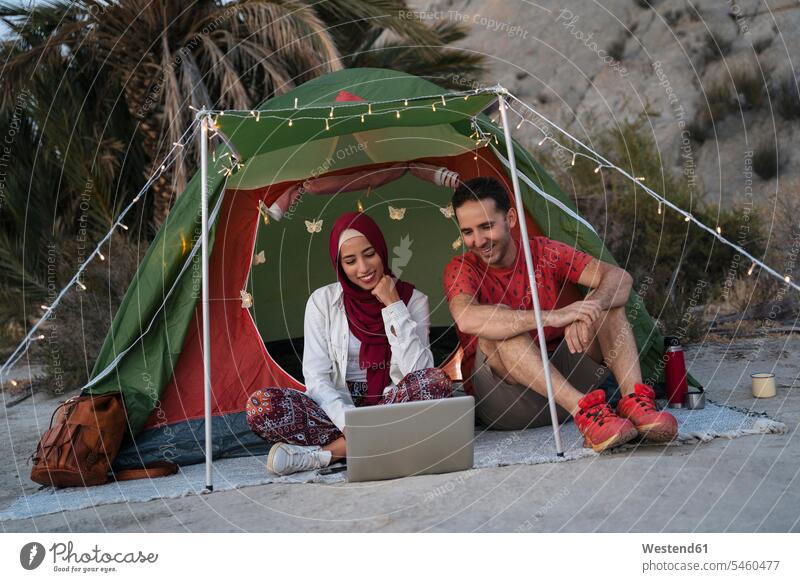 Junge Frau mit Hijab und Mann mit Laptop in einem Zelt Kopftücher Rechner Laptops Notebook Notebooks verreisen sitzend sitzt abends freuen Campen zelten mobil
