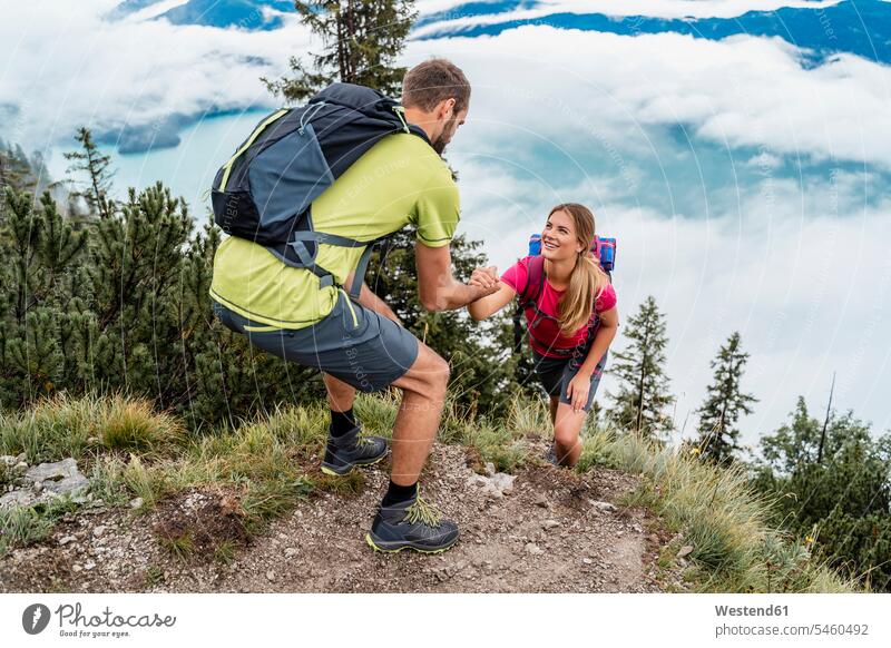Junger Mann hilft seiner Freundin bei einer Wanderung in den Bergen, Herzogstand, Bayern, Deutschland Touristen Rucksäcke T-Shirts gehend geht Griff steigen