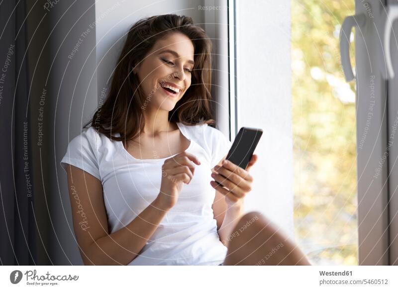 Junge Frau sitzt auf dem Fensterbrett und benutzt ein Smartphone sitzen sitzend junge Frau junge Frauen Zuhause zu Hause daheim Fensterbank Fenstersims