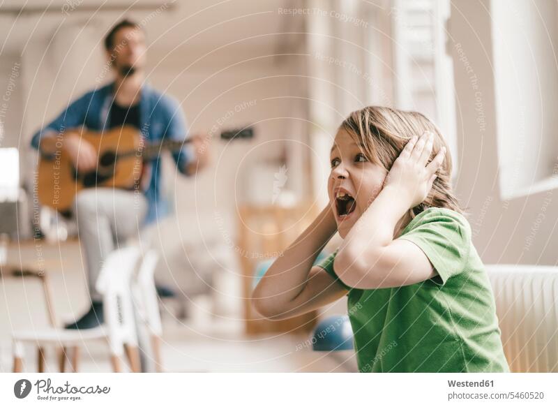 Entsetzter Sohn hält sich die Ohren zu, während der Vater zu Hause Gitarre spielt musizieren Musik machen Gitarren Söhne Zuhause daheim Papas Väter Vati Vatis
