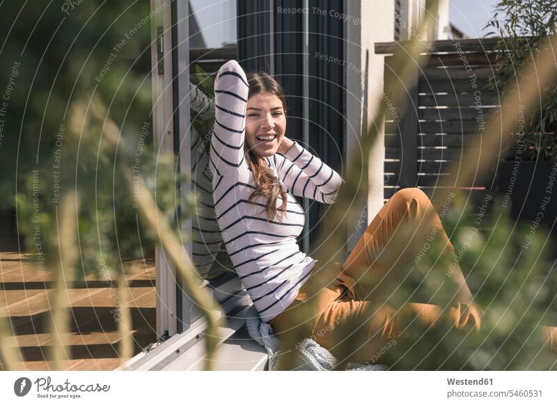 Porträt einer lachenden jungen Frau, die zu Hause auf der Terrasse sitzt Deutschland bequem Hand hinter dem Kopf Hände hinter dem Kopf Haende hinter dem Kopf