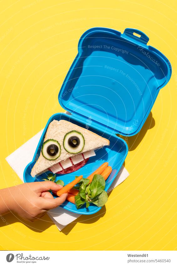 Hand eines kleinen Mädchens, das eine Babykarotte aus einer Lunchbox mit einem lustig aussehenden Sandwich mit anthropomorphem Gesicht aufhebt Draufsicht