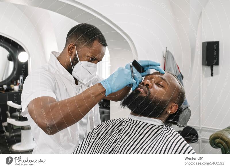 Mann mit Vollbart wird rasiert, Barbier trägt chirurgische Maske und Handschuhe Leute Menschen People Person Personen Kunden Kundschaft Job Berufe