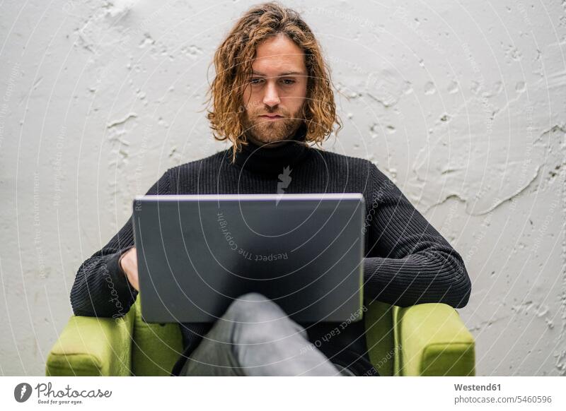 Bärtiger junger Mann mit Laptop, während er zu Hause auf einem Sessel an der Wand sitzt Farbaufnahme Farbe Farbfoto Farbphoto Spanien Freizeitbeschäftigung Muße