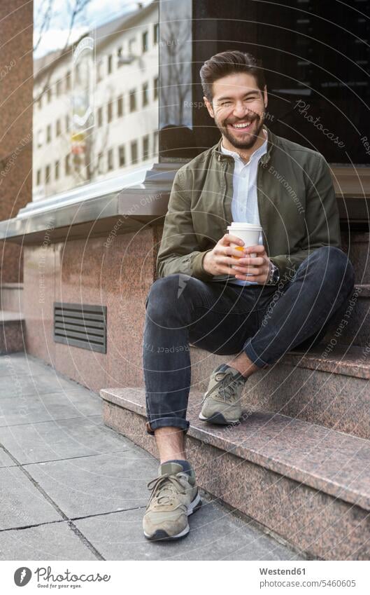 Porträt eines lachenden jungen Mannes auf einer Treppe sitzend mit Kaffee zum Mitnehmen sitzt freuen Muße Spass spassig spaßig Spässe Späße außen draußen