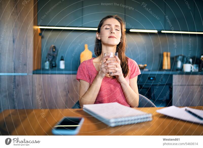 Lächelnde Unternehmerin mit geschlossenen Augen hält Trinkglas am Schreibtisch im Heimbüro Farbaufnahme Farbe Farbfoto Farbphoto Freizeitkleidung