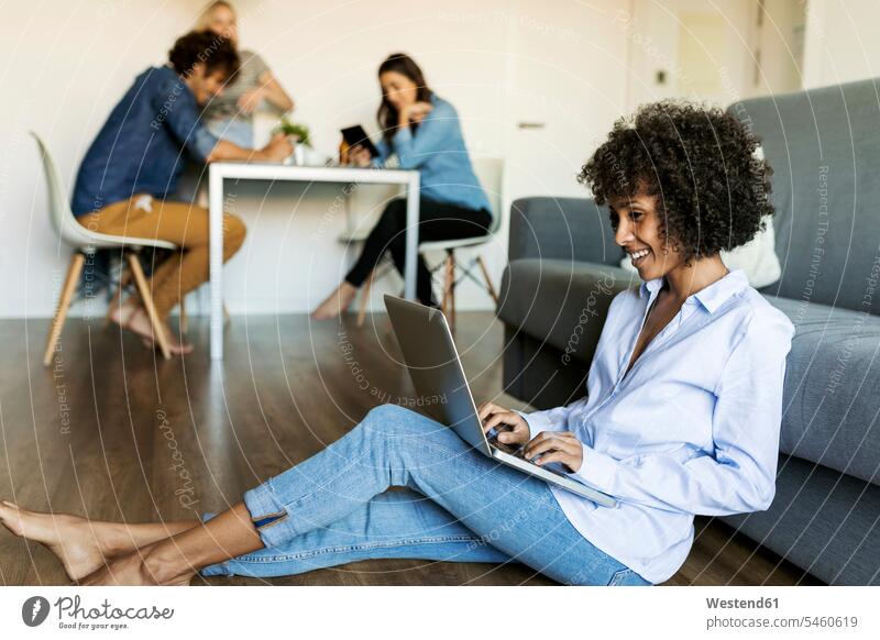 Lächelnde Frau sitzt auf dem Boden mit Laptop mit Freunden im Hintergrund Böden Boeden Notebook Laptops Notebooks lächeln weiblich Frauen sitzen sitzend