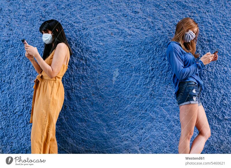Maskierte Freundinnen mit Smartphones, die an der blauen Mauer in der Stadt stehen Farbaufnahme Farbe Farbfoto Farbphoto Spanien Freizeitbeschäftigung Muße Zeit
