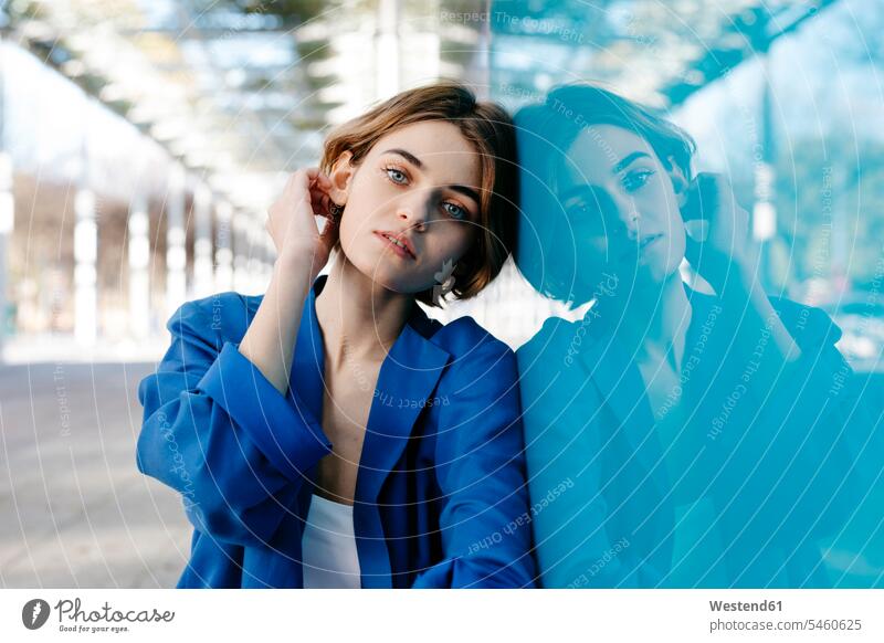 Junge Frau mit urbanem Aussehen sitzt auf dem Boden und lehnt mit ihrem Spiegelbild an einer bunten Glaswand Glasscheiben Hosen Jacken sitzend blaue blauer
