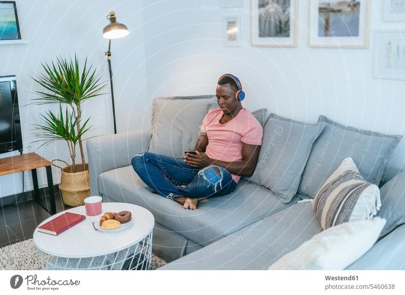 Junger Mann mit Kopfhörer und Smartphone auf der Couch zu Hause Leute Menschen People Person Personen Afrikanisch Afrikanische Abstammung dunkelhäutig Farbige