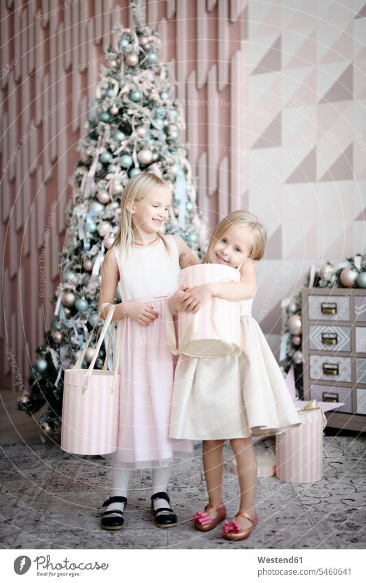 Porträt von zwei fröhlichen kleinen Mädchen, die vor einem beleuchteten Weihnachtsbaum mit Geschenkkartons stehen Leute Menschen People Person Personen