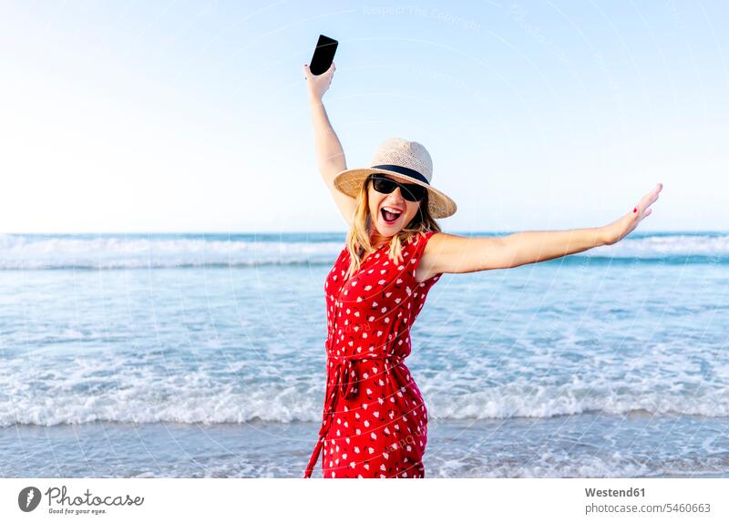 Lächelnde blonde Frau in rotem Kleid und Hut mit Smartphone in der Hand am Strand Telekommunikation telefonieren Handies Handys Mobiltelefon Mobiltelefone