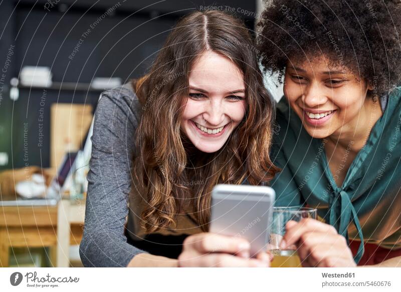 Zwei lächelnde junge Frauen teilen sich ein Mobiltelefon im Büro Handy Handies Handys Mobiltelefone Teilen Sharing weiblich Office Büros Telefon telefonieren