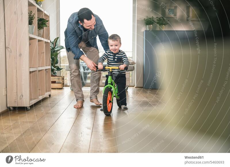 Vater hilft Sohn beim Fahren mit einem Balance-Fahrrad zu Hause Bikes Fahrräder Räder Rad fahren lehren unterrichten beibringen Zuhause daheim Kinderlaufrad