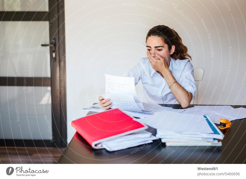 Schockierte Studentin liest Dokument am Schreibtisch zu Hause Tische Arbeitstisch Schreibtische Papiere Lektüre sitzend sitzt pessimistisch schwarzseherisch