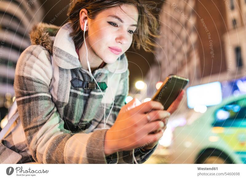 Spanien, Madrid, junge Frau in der Stadt bei Nacht, die ihr Smartphone benutzt und Kopfhörer trägt nachts Ohrhörer iPhone Smartphones weiblich Frauen staedtisch