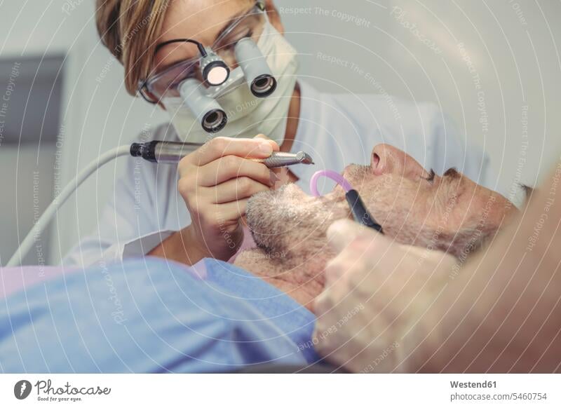 Patient bekommt zahnärztliche Behandlung, Zahnarzt mit Zahnbohrer und Kopf Lupen und Licht Kranke Kranker Patienten Assistent Assistenten Stirnlupe Lupenbrille