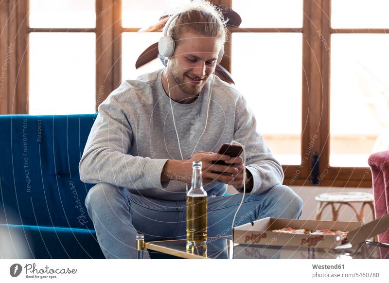 Lächelnder junger Mann sitzt auf Couch mit Handy und Kopfhörer Leute Menschen People Person Personen Alleinstehende Alleinstehender Singles Unverheiratete