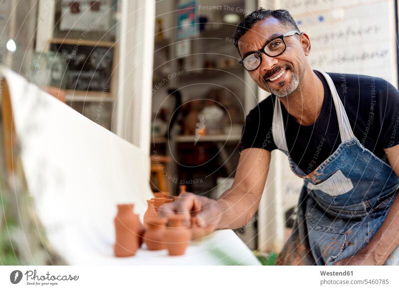 Lächelnder männlicher Töpfer mit Miniaturen auf Tisch bei der Arbeit in der Werkstatt Farbaufnahme Farbe Farbfoto Farbphoto Spanien Innenaufnahme Innenaufnahmen
