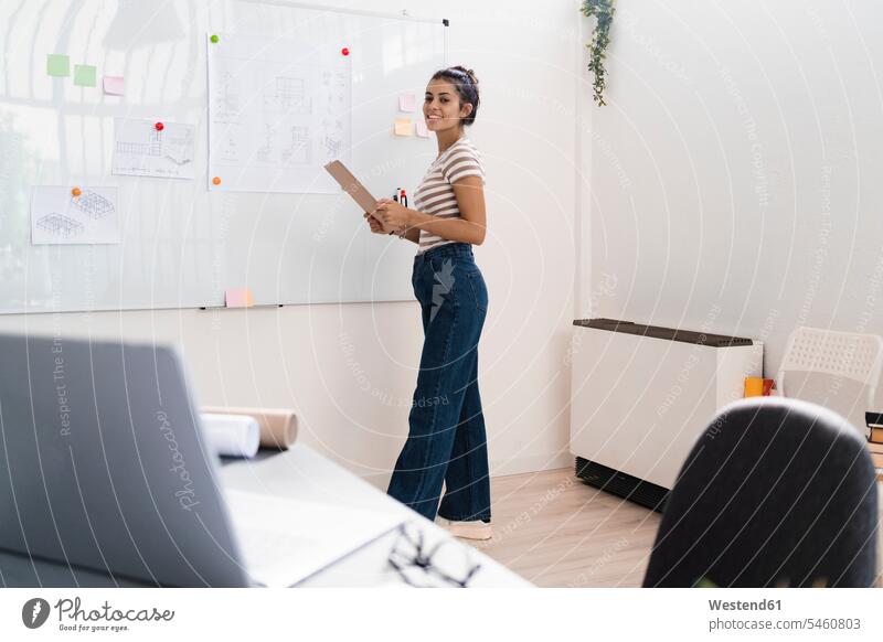 Lächelnde, schöne, junge Architektin hält Klemmbrett, während sie am Whiteboard am kreativen Arbeitsplatz steht Farbaufnahme Farbe Farbfoto Farbphoto