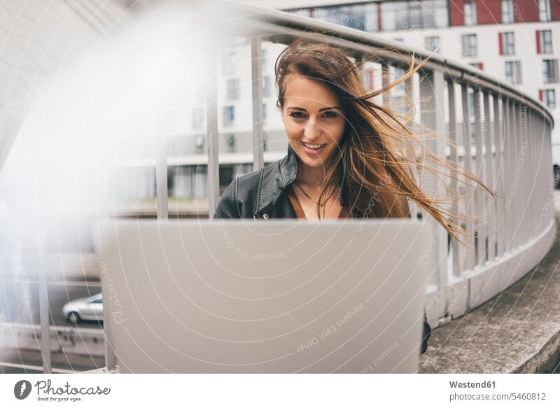 Porträt einer lächelnden jungen Frau mit windgepeitschtem Haar mit Laptop auf der Autobahnbrücke Autobahnen Notebook Laptops Notebooks weiblich Frauen Haare