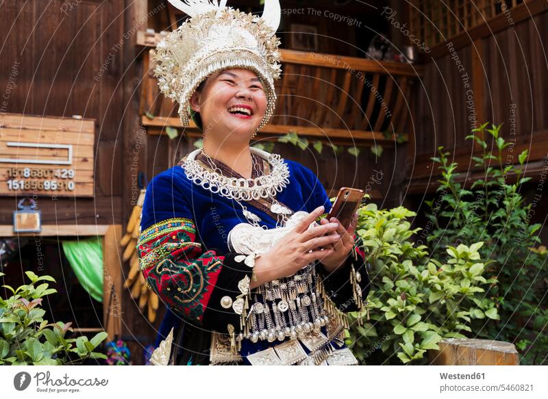 China, Guizhou, lachende Miao-Frau mit traditionellem Kleid und Kopfschmuck, die ein Mobiltelefon hält Smartphone iPhone Smartphones festlich Kopfbedeckung