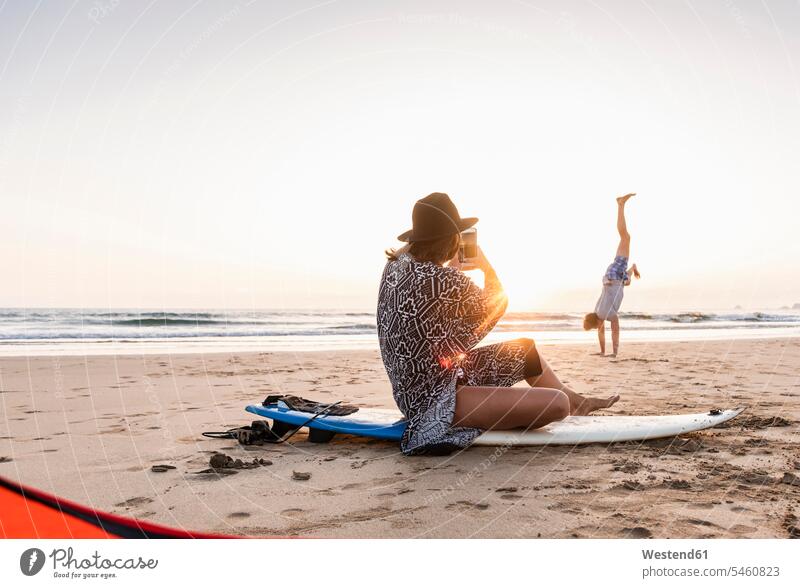 Junge Frau sitzt auf einem Surfbrett und fotografiert einen jungen Mann, der am Strand Handstände übt Handstand Handstaende benutzen benützen üben ausüben Übung