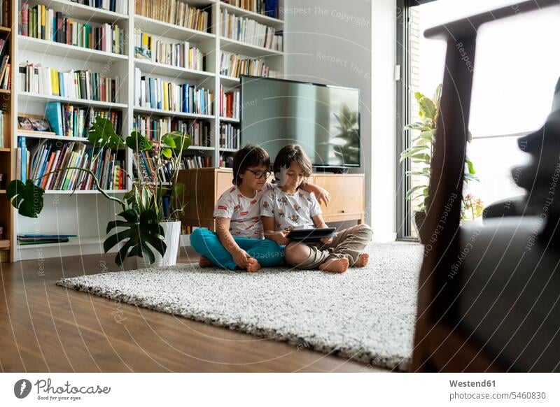 Zwei Brüder sitzen mit einem digitalen Tablett auf dem Boden im Wohnzimmer Leute Menschen People Person Personen Europäisch Kaukasier kaukasisch sehen sehend