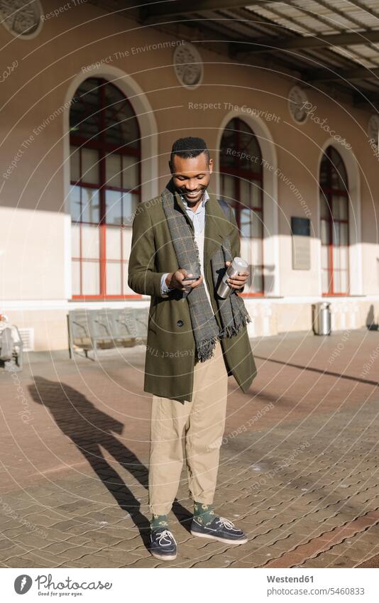 Stylischer Mann mit wiederverwendbarem Becher, der während des Wartens auf den Zug ein Smartphone benutzt geschäftlich Geschäftsleben Geschäftswelt