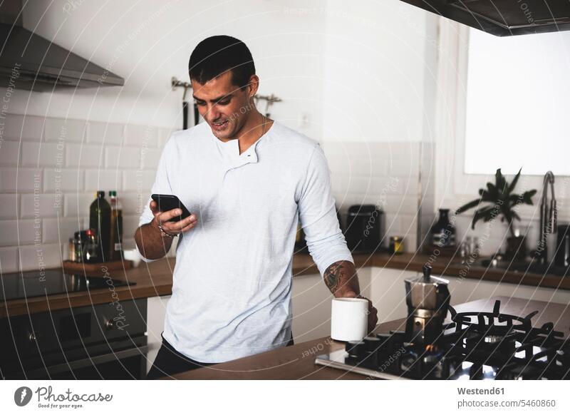 Lächelnder junger Mann mit Tasse Kaffee mit Handy in der Küche zu Hause lächeln Kaffeetasse Kaffeetassen Männer männlich Zuhause daheim Mobiltelefon Handies