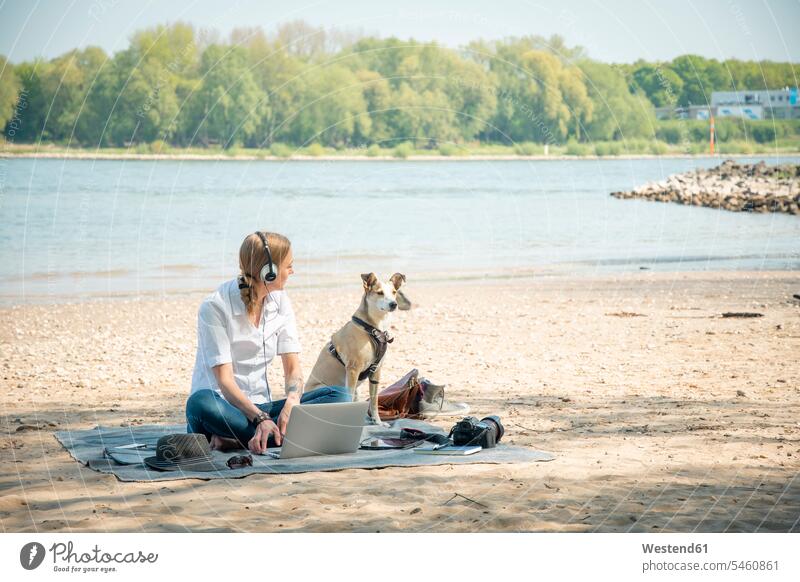 Frau sitzt auf einer Decke an einem Fluss mit Hund, trägt Kopfhörer und benutzt einen Laptop Notebook Laptops Notebooks Decken Hunde weiblich Frauen sitzen