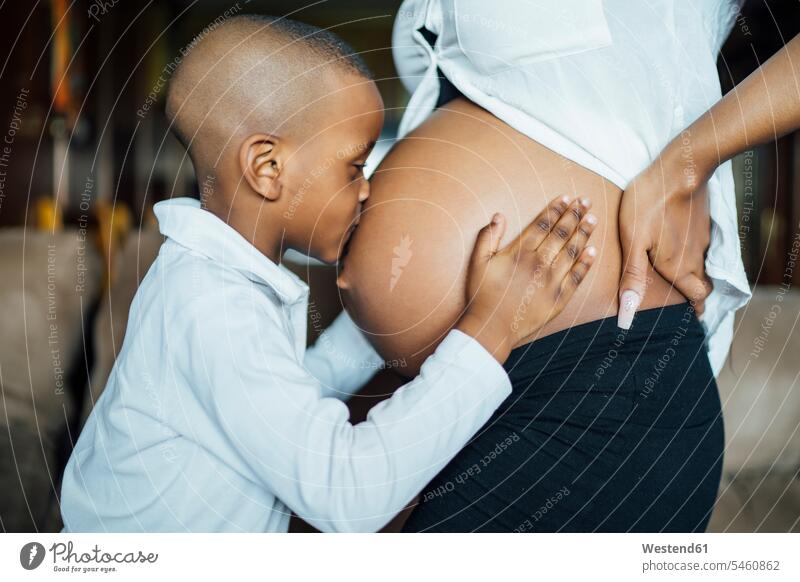 Kleiner Junge küsst den Bauch einer schwangeren jungen Frau anfassen Berührung Kuss Küsse freuen zufrieden stehend steht daheim zu Hause Schwangere