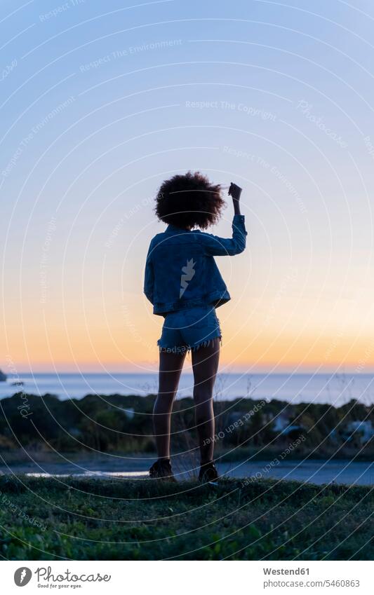 Rückansicht einer jungen Frau mit Hand im Haar, die bei Sonnenuntergang auf einem Aussichtspunkt steht, Ibiza abends zufrieden stehend frei Muße Individuell