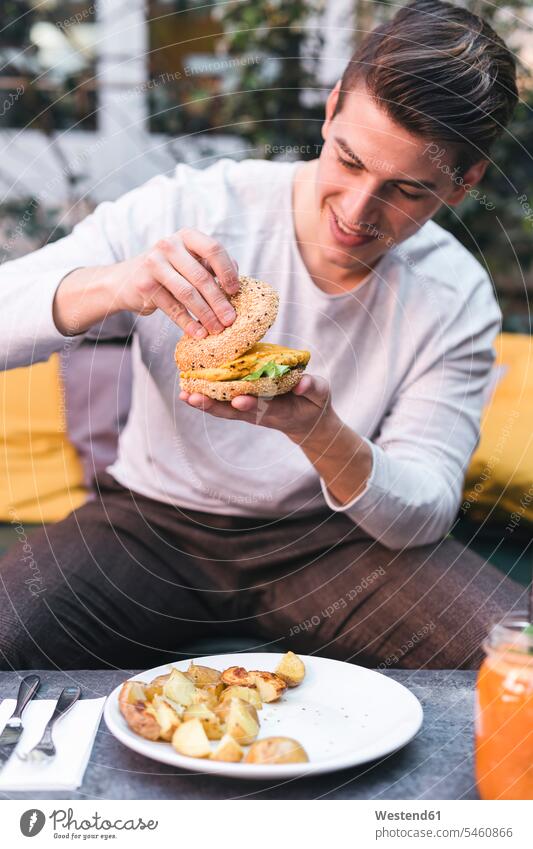 Junger Mann sitzt auf der Couch in einem Restaurant und isst einen veganen Burger zum Mittagessen Model Modell Modelle Models Freizeitkleidung casual