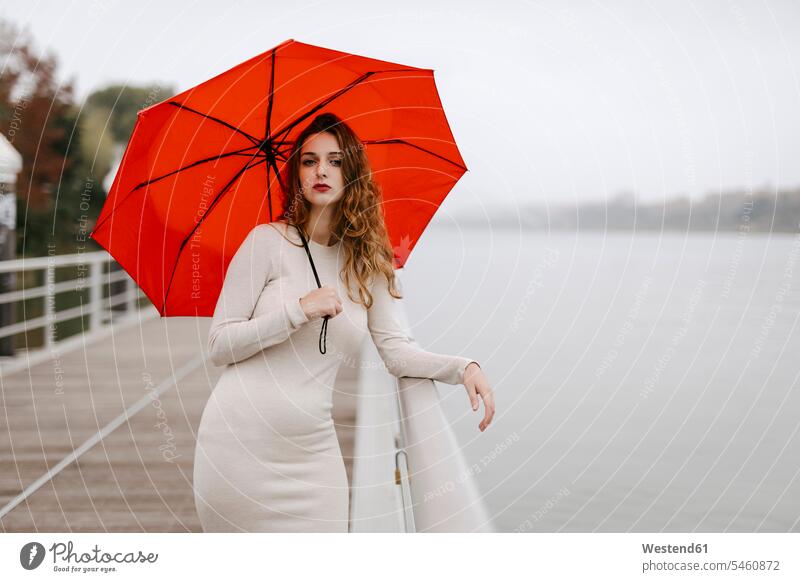 Porträt einer jungen Frau mit rotem Regenschirm, die sich an einem Regentag an ein Geländer lehnt Kleider Schirme Regenschirme Erwartung sehnsüchtig Streben