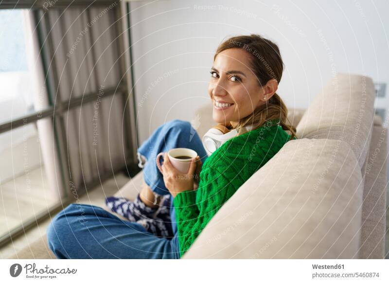 Frau trinkt Kaffee, während sie zu Hause auf dem Sofa sitzt Farbaufnahme Farbe Farbfoto Farbphoto Innenaufnahme Innenaufnahmen innen drinnen Tag