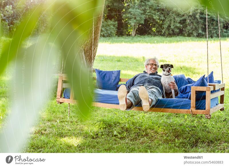 Senior Mann entspannt auf einem Schaukelbett in seinem Garten mit seinem Hund Freundschaft Freunde Kameradschaft Gärten Gaerten Entspannung Entspannen relaxen
