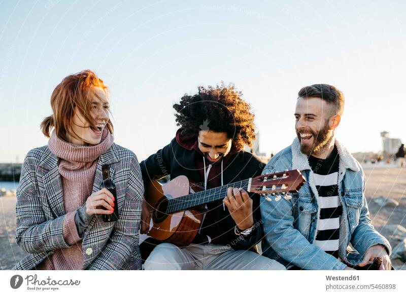 Drei glückliche Freunde mit Gitarre sitzen im Freien bei Sonnenuntergang Glück glücklich sein glücklichsein Gitarren sitzend sitzt Sonnenuntergänge Freundschaft